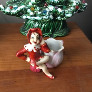 Könnte beinhalten: Eine Keramikfigur eines rot gekleideten Elfen, der auf einem rosafarbenen Blumentopf sitzt. Der Elf hat einen weißen Kragen und weiße Punkte auf dem roten Kleid. Der Elf hält sich eine Hand an die Wange.