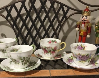 May include: Four floral teacups and saucers with delicate floral patterns. The cups are white with green, pink, and purple flowers. The saucers have a matching floral design with a green rim.