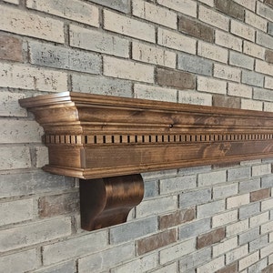 May include: A dark brown wooden mantel shelf with a decorative trim and a curved bracket on the left side. The shelf is mounted on a white brick wall.