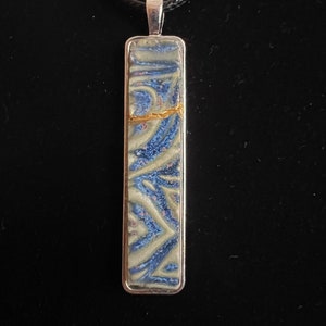 Pode incluir: Um colar com um pingente retangular de vidro azul e branco com detalhes dourados. O pingente tem um padrão de redemoinho.