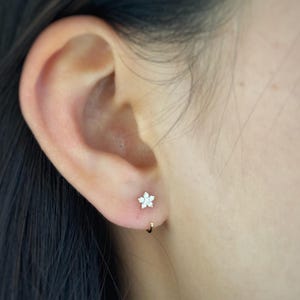 Puede incluir: Primer plano de una oreja con un pequeño pendiente de aro dorado y un pendiente de botón en forma de flor. El pendiente de botón tiene un centro blanco y pétalos blancos. La joya es delicada.