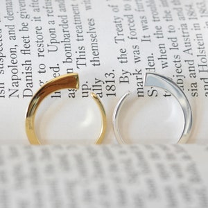 May include: Two simple, minimalist rings. One ring is gold and the other is silver. Both rings are open and have a slightly angled design.