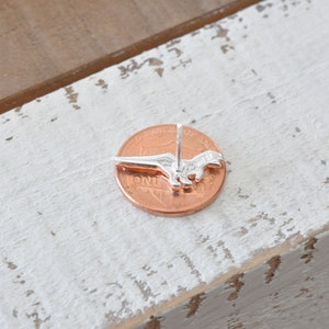 May include: Silver dinosaur earring stud on a penny for size comparison. The earring is a simple design with a detailed dinosaur silhouette.