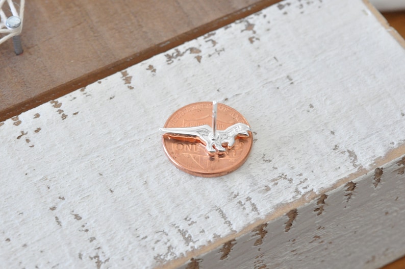 May include: Silver dinosaur earring stud on a penny for size comparison. The earring is a simple design with a detailed dinosaur silhouette.