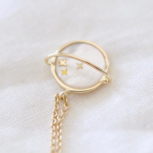 Könnte beinhalten: Eine goldene Halskette mit einem runden Anhänger, der mit einer weißen Perlmutteinlage und vier kleinen goldenen Sternen verziert ist. Der Anhänger ist an einer zarten goldenen Kette befestigt.