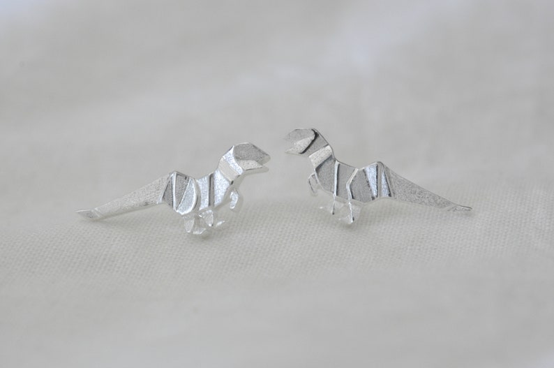 May include: A pair of silver origami dinosaur earrings. The dinosaurs are facing each other.