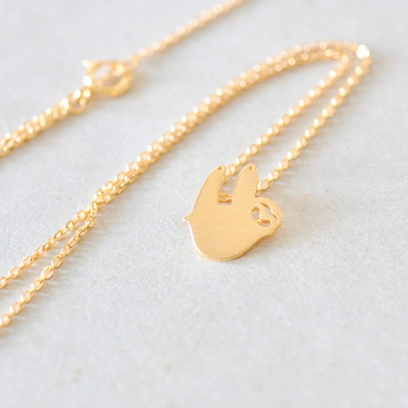 K&ouml;nnte beinhalten: Eine goldfarbene Halskette mit einem Faultier-Anh&auml;nger. Der Faultier-Anh&auml;nger ist eine Vollsilhouette mit einem kleinen Ausschnitt. Die Kette ist filigran und etwa 45 cm lang. Die Halskette wird auf einer hellen Oberfl&auml;che pr&auml;sentiert.