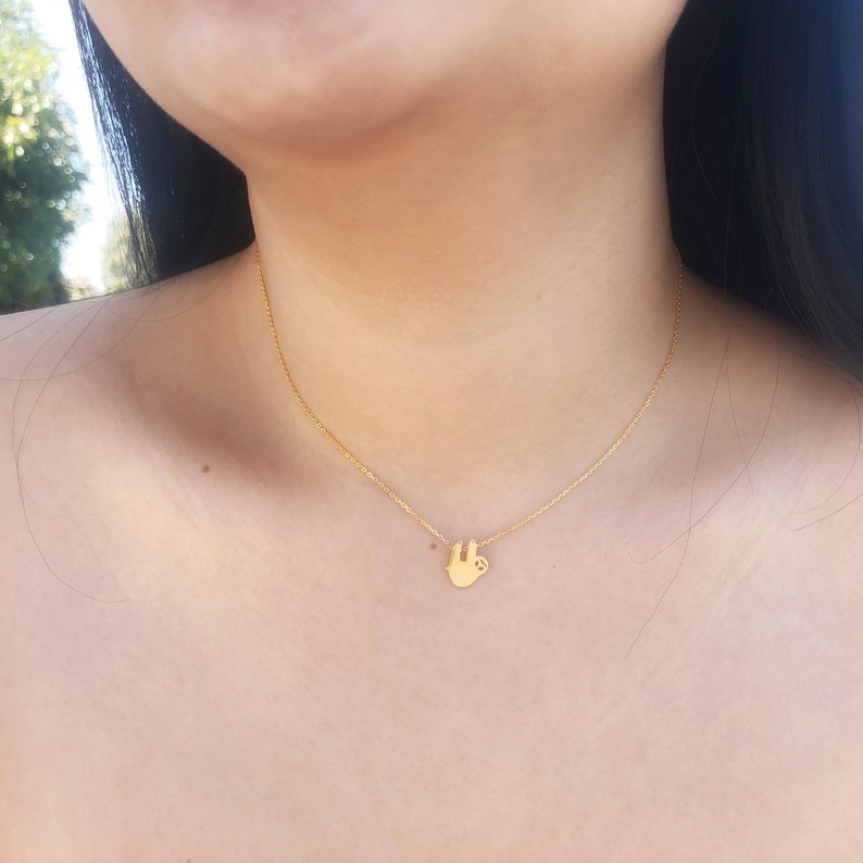 K&ouml;nnte beinhalten: Eine zarte, goldfarbene Halskette mit einem kleinen Faultier-Anh&auml;nger. Das Faultier ist in h&auml;ngender Pose mit ausgestreckten Armen dargestellt. Die Halskette ist d&uuml;nn und etwa 45 cm lang.