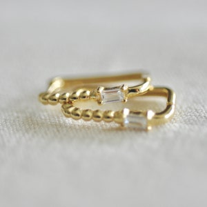 May include: Two gold hoop earrings with a single baguette diamond set in each hoop. The hoops are beaded and have a rounded shape.