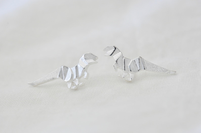 May include: A pair of silver origami dinosaur earrings. The dinosaurs are facing opposite directions.