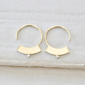 May include: A pair of gold-colored hoop earrings. Each earring features a curved, open hoop design with a flat, rectangular base. A small, clear gemstone is set in the center of the base. The earrings are displayed on a neutral-colored surface.