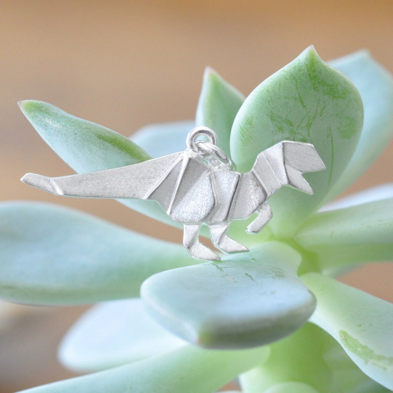 May include: Silver origami dinosaur charm pendant. The charm is silver and has a detailed origami design. The charm is sitting on a green succulent plant.