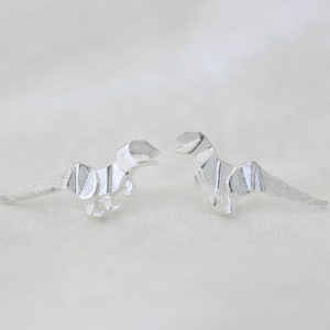 May include: A pair of silver origami dinosaur earrings. The dinosaurs are facing each other.