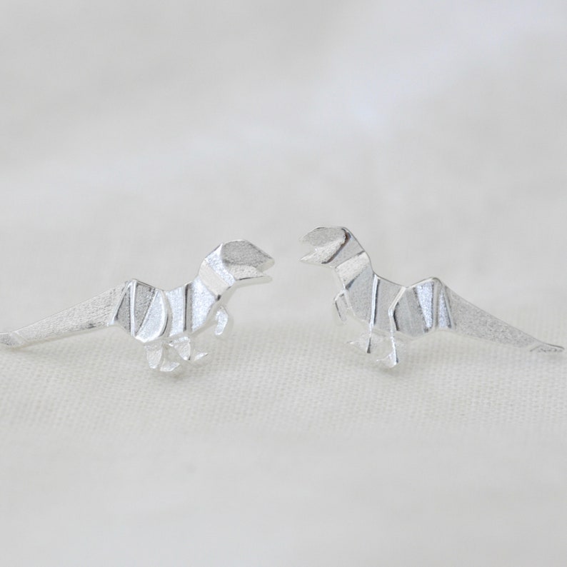 May include: A pair of silver origami dinosaur earrings. The dinosaurs are facing each other.
