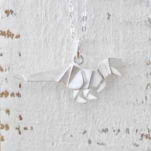 May include: Silver origami dinosaur pendant necklace. The pendant is a detailed, three-dimensional representation of a dinosaur folded in the origami style.