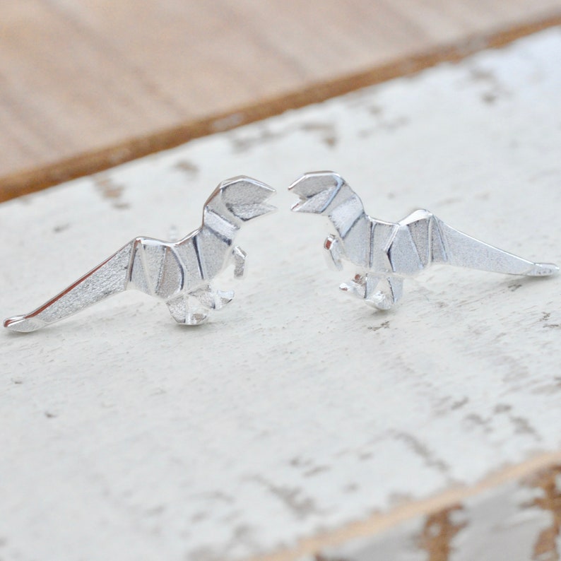 May include: A pair of silver origami dinosaur earrings. The dinosaurs are facing each other and have a detailed, folded design.