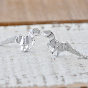 May include: A pair of silver origami dinosaur earrings. The dinosaurs are facing each other and are made of a textured metal.
