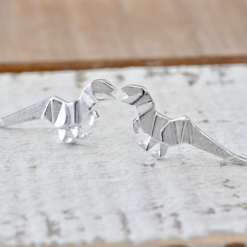May include: A pair of silver origami dinosaur earrings. The dinosaurs are facing each other and are made of a textured metal.