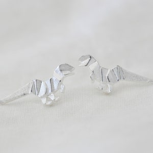 May include: A pair of silver origami dinosaur earrings. The dinosaurs are facing each other and have a textured surface.
