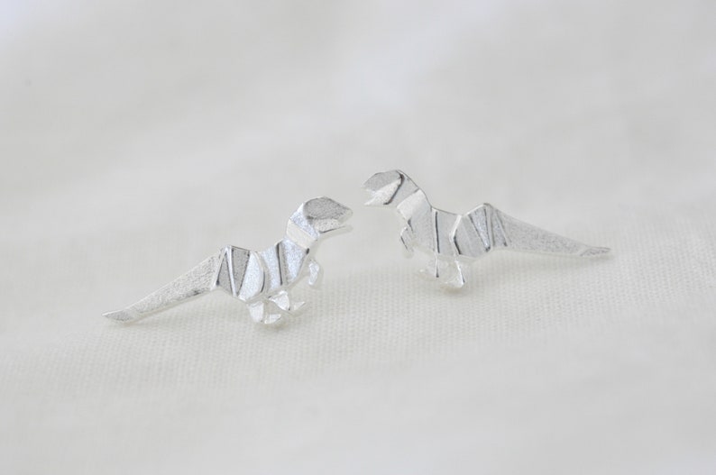 May include: A pair of silver origami dinosaur earrings. The dinosaurs are facing each other and have a textured surface.