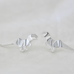 May include: A pair of silver origami dinosaur earrings. The dinosaurs are facing each other and have a detailed, folded design.