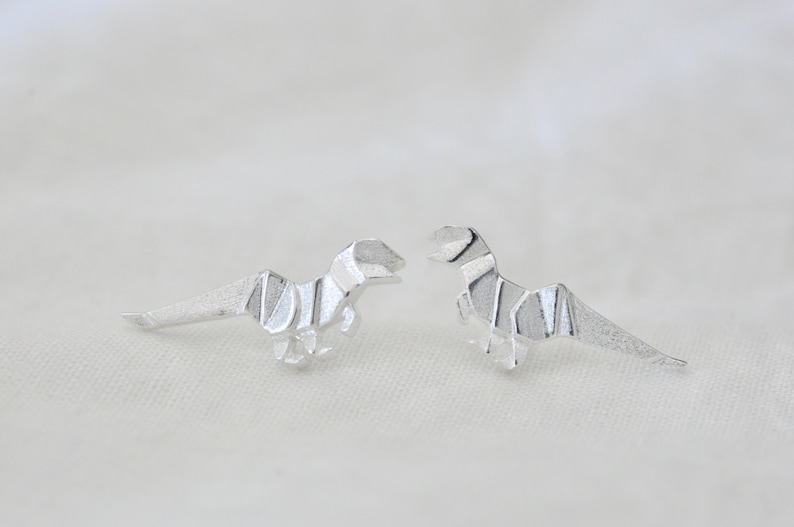 May include: A pair of silver origami dinosaur earrings. The dinosaurs are facing each other and have a detailed, folded design.