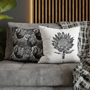 May include: Two decorative throw pillows on a gray couch. One pillow features a black and white floral pattern, while the other pillow has a single black and white illustration of a protea flower.