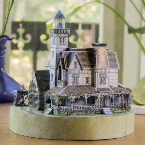 Op de afbeelding: Een miniatuur papieren model van een huis met meerdere verdiepingen, een hoge toren, een grijs pannendak en een veranda. De structuur heeft gedetailleerde witte gevels, donkere raamkozijnen en een stenen gedeelte, geplaatst op een ronde voet.