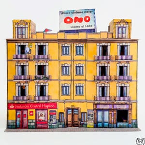 Op de afbeelding: Papieren model van een geel gebouw met de tekst "DESENGAÑO, 21" in het geel. Het gebouw heeft meerdere ramen, balkons en etalages, waaronder een Santander Central Hispano. Het model heeft ook een ONO-bord.