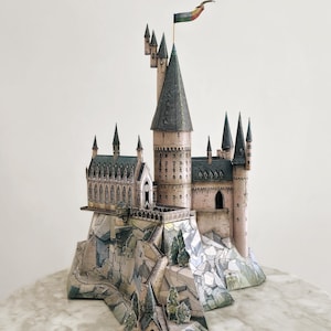 Può includere: Un modello di carta dettagliato di un castello con più torri, un'alta guglia centrale e una bandiera colorata. Il castello si trova su una base rocciosa con un ponte e piccoli alberi. Il modello è beige, grigio e verde.