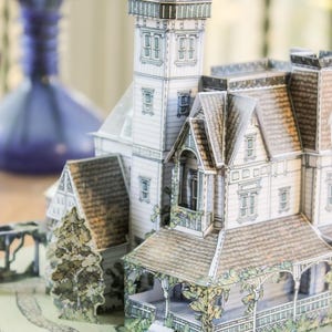 Op de afbeelding: Een papieren model van een huis in victoriaanse stijl met een aangebouwde vuurtoren. De structuur heeft witte gevelbekleding, bruine pannendaken en groene accenten van gebladerte. Het staat op een ronde basis met een stenen pad en boompjes.