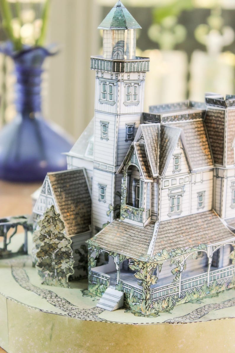 Op de afbeelding: Een papieren model van een huis in victoriaanse stijl met een aangebouwde vuurtoren. De structuur heeft witte gevelbekleding, bruine pannendaken en groene accenten van gebladerte. Het staat op een ronde basis met een stenen pad en boompjes.
