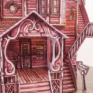 May include: A pink paper model of a house with a porch, stairs, and a sign that says "Barnyard". The house has a number "22990" and the words "Apt A", "Apt B", and "Apt C" on the side.