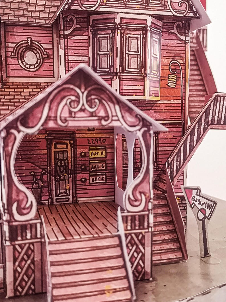 May include: A pink paper model of a house with a porch, stairs, and a sign that says "Barnyard". The house has a number "22990" and the words "Apt A", "Apt B", and "Apt C" on the side.