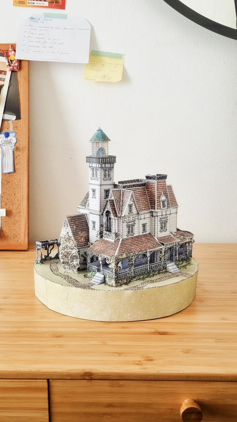 Op de afbeelding: Een gedetailleerd papieren model van een huis in victoriaanse stijl met een aangebouwde vuurtoren. De structuur heeft witte muren, roodbruine pannendaken, meerdere ramen en een veranda, geplaatst op een ronde ondergrond.