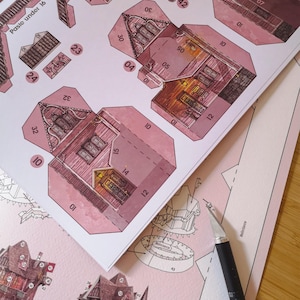 May include: A pink papercraft model of a house with detailed illustrations and numbered tabs for assembly. The model is partially assembled and shows the interior of the house with windows and a fireplace. The text "Paste under 16" and "Paste under" are visible on the paper.