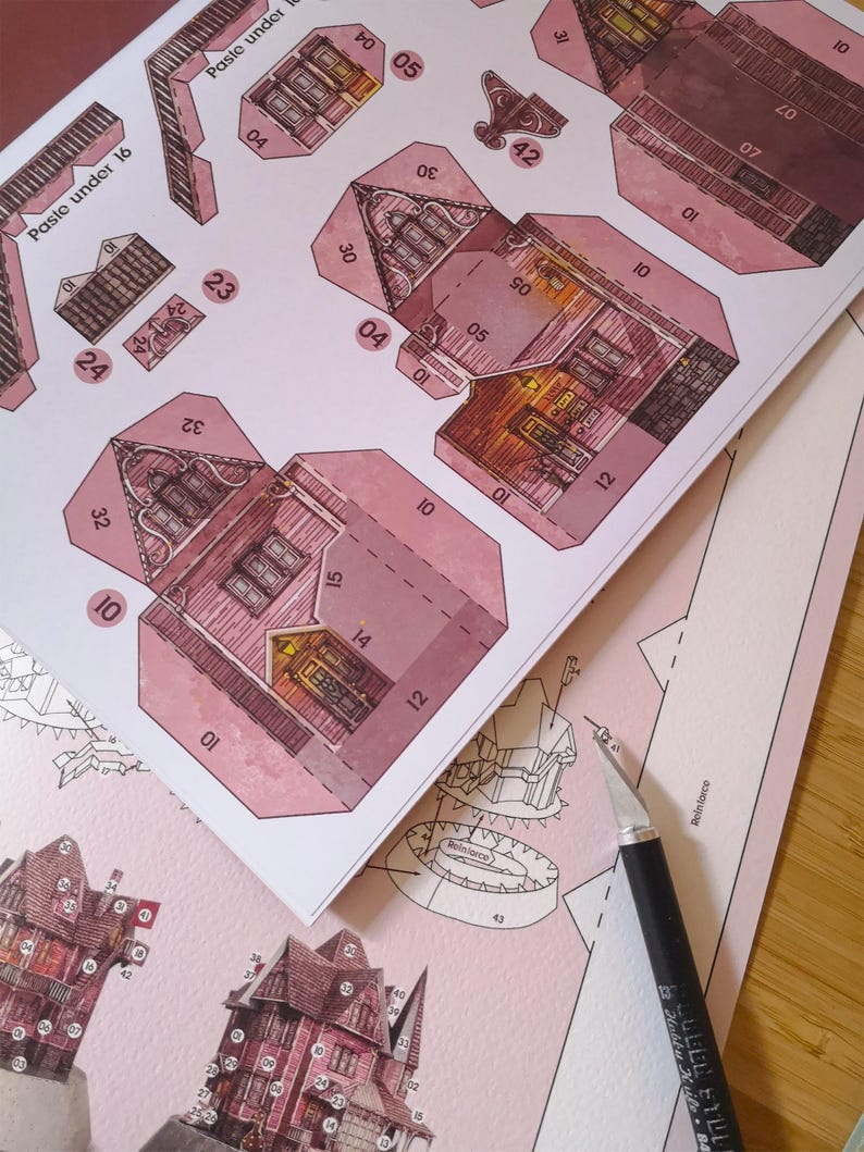 May include: A pink papercraft model of a house with detailed illustrations and numbered tabs for assembly. The model is partially assembled and shows the interior of the house with windows and a fireplace. The text "Paste under 16" and "Paste under" are visible on the paper.