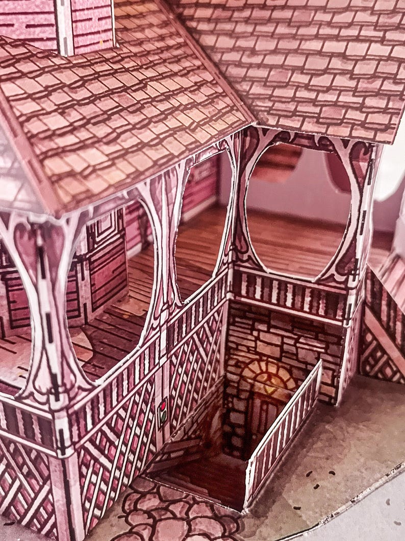 May include: A paper model of a pink and brown house with a detailed porch and a stone basement. The house has a gabled roof with a shingled pattern and a decorative railing around the porch.