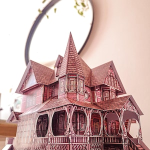 May include: A detailed paper model of a Victorian-style house with a pointed roof, intricate trim, and a wraparound porch. The house is painted in shades of pink and white.