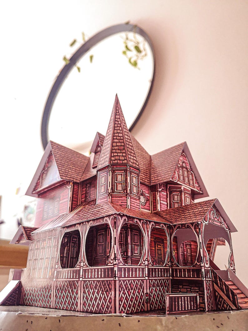 May include: A detailed paper model of a Victorian-style house with a pointed roof, intricate trim, and a wraparound porch. The house is painted in shades of pink and white.