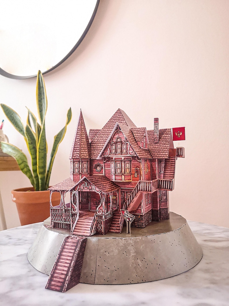May include: A paper model of a red Victorian house with a porch and a flag on top. The house is sitting on a gray base.