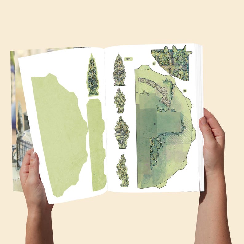 Op de afbeelding: Een papieren knutselboek open op een pagina met gedrukte uitknipvormen voor een miniatuurtuinsc&egrave;ne. De pagina toont lichtgroene landschapsdelen, diverse boomillustraties en een stenen padontwerp met stippellijnen om uit te knippen.