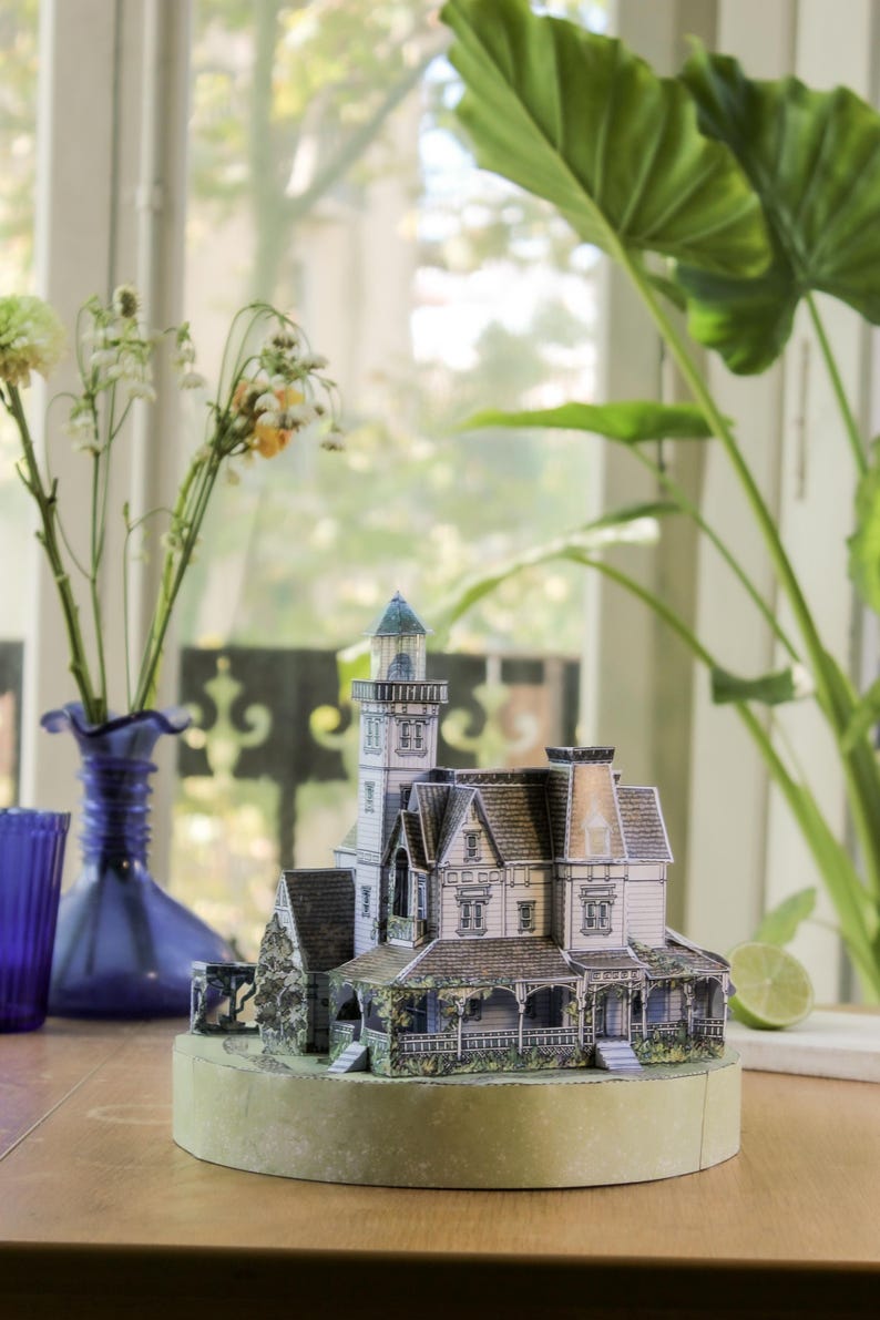 Op de afbeelding: Een miniatuur papieren model van een huis met meerdere verdiepingen, een hoge toren, een grijs pannendak en een veranda. De structuur heeft gedetailleerde witte gevels, donkere raamkozijnen en een stenen gedeelte, geplaatst op een ronde voet.