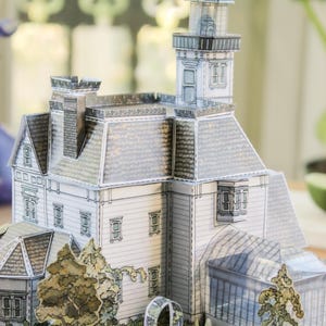 Op de afbeelding: Een papieren knutselmodel van een huis met meerdere verdiepingen, voorzien van een hoge toren, een schoorsteen en een glazen serre. Het bouwwerk heeft witte gevels, een grijs pannendak en is omringd door papieren uitsparingen van bomen en een tuinboog.