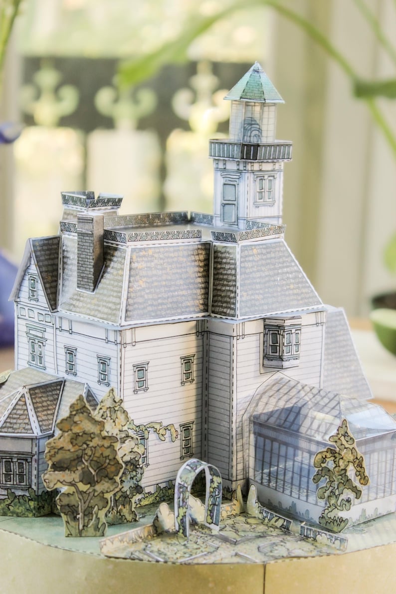 Op de afbeelding: Een papieren knutselmodel van een huis met meerdere verdiepingen, voorzien van een hoge toren, een schoorsteen en een glazen serre. Het bouwwerk heeft witte gevels, een grijs pannendak en is omringd door papieren uitsparingen van bomen en een tuinboog.