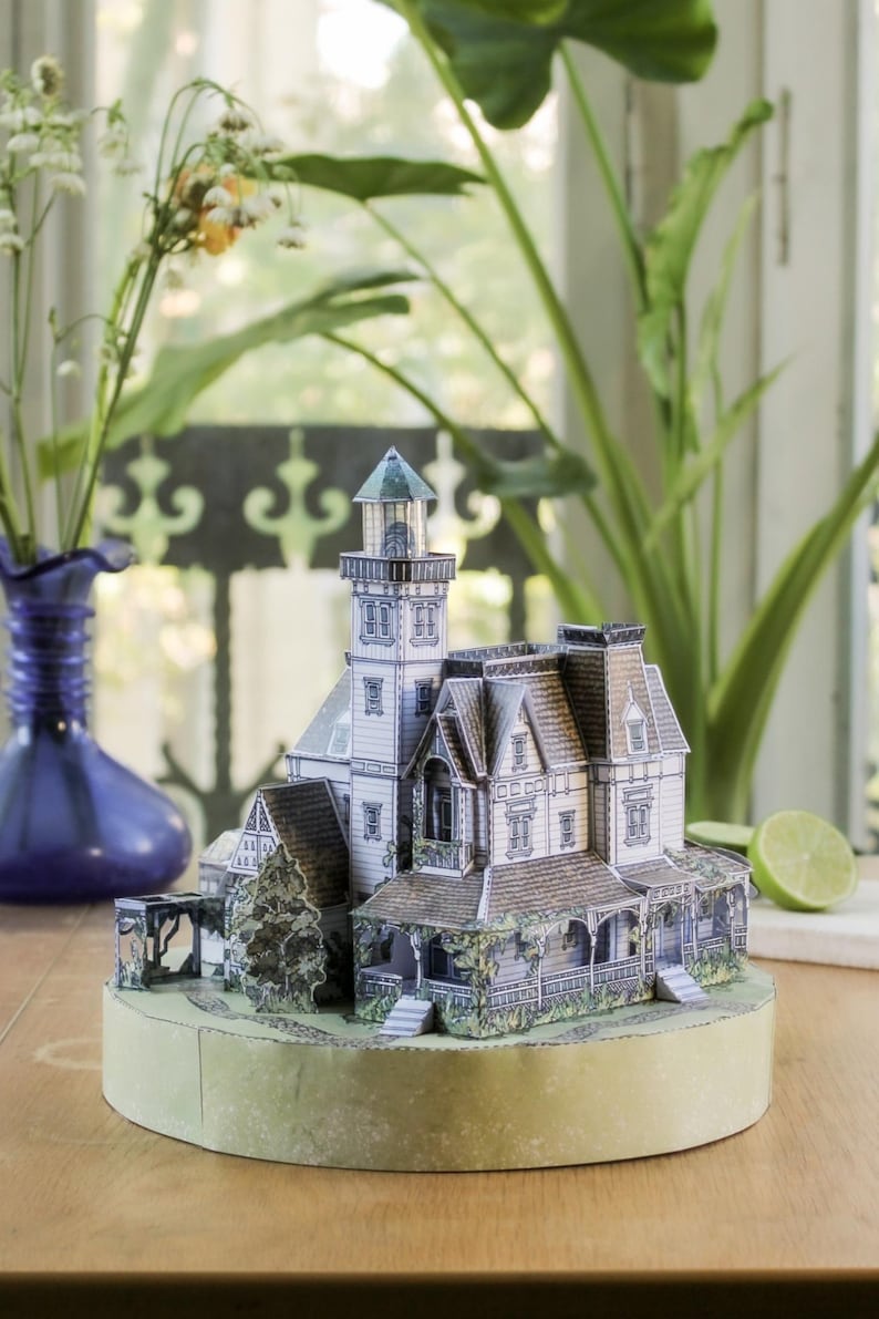 Op de afbeelding: Een papieren model van een huis in victoriaanse stijl met een aangebouwde vuurtoren, voorzien van gedetailleerde prints van dakspanen, ramen en veranda's. De structuur is gemonteerd op een ronde, lichtgroene basis met paden.
