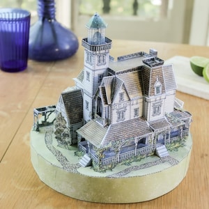 Op de afbeelding: Een driedimensionaal papieren model van een huis in victoriaanse stijl met een aangebouwde vuurtoren, voorzien van gedetailleerde prints van pannendaken, veranda's en stenen paden, gemonteerd op een ronde lichtgroene basis.