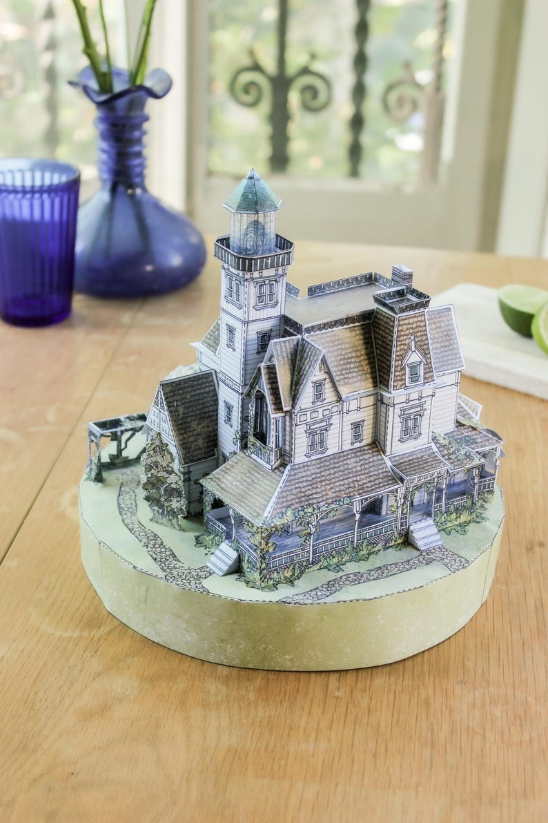 Op de afbeelding: Een driedimensionaal papieren model van een huis in victoriaanse stijl met een aangebouwde vuurtoren, voorzien van gedetailleerde prints van pannendaken, veranda's en stenen paden, gemonteerd op een ronde lichtgroene basis.