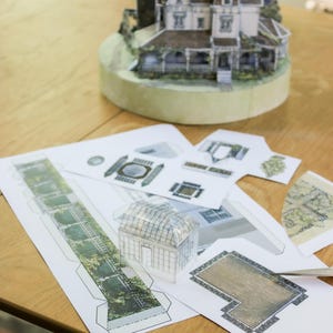Op de afbeelding: Een papieren knutselset voor een miniatuurhuismodel, met bedrukte vellen met architecturale onderdelen, een gedeeltelijk geassembleerde kasstructuur en een voltooid driedimensionaal huismodel op een ronde basis.