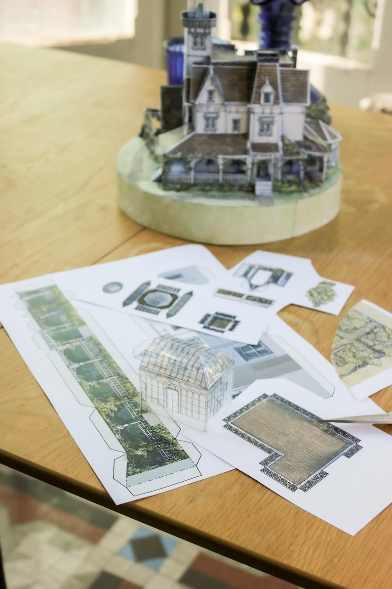 Op de afbeelding: Een papieren knutselset voor een miniatuurhuismodel, met bedrukte vellen met architecturale onderdelen, een gedeeltelijk geassembleerde kasstructuur en een voltooid driedimensionaal huismodel op een ronde basis.